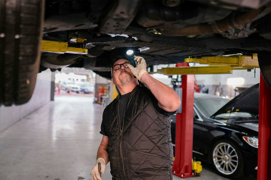 Auto mechanic using a flashlight to inspect the underside of a car elevated on a LOADED two post lift in a workshop.